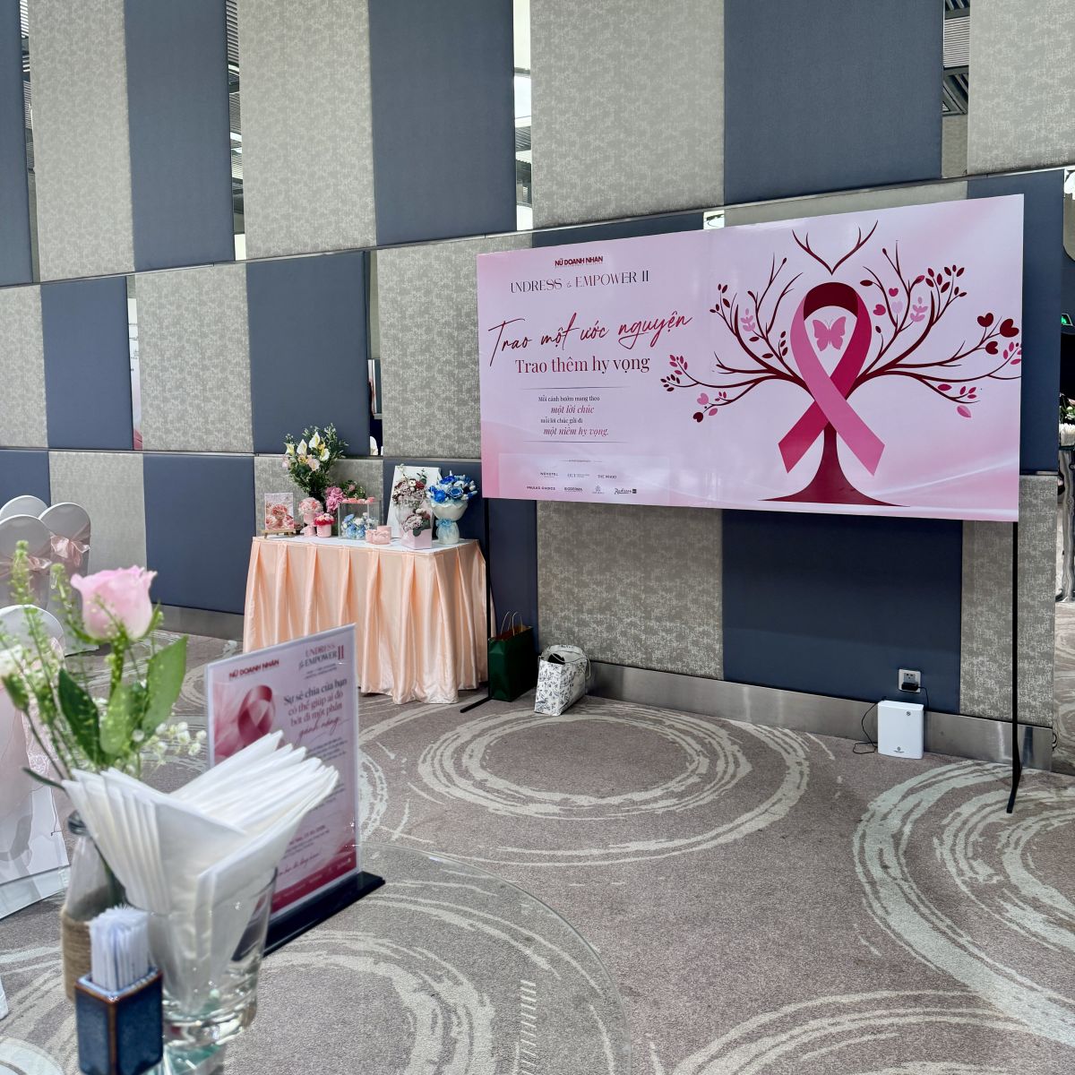 Backdrop thông điệp Undress to Empower II tông hồng bên khu vực trưng bày hoa.