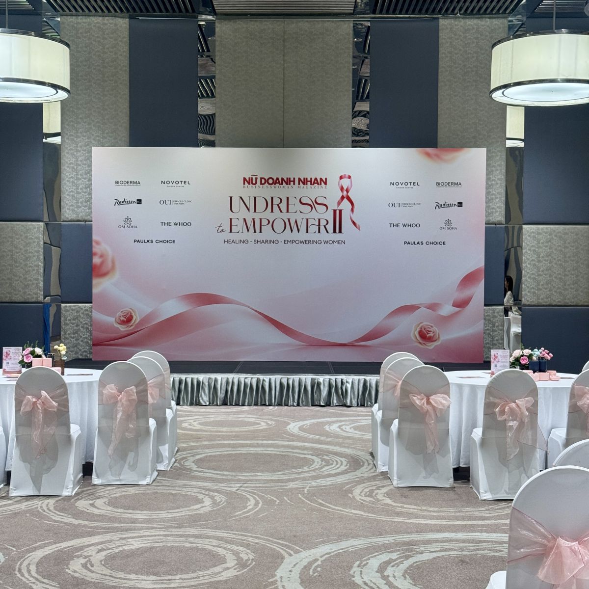 Backdrop sân khấu Undress to Empower II với dải ruy băng hồng và nền trắng.